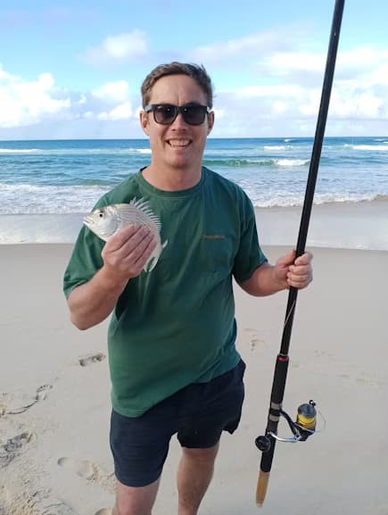 Dan on a beach, holding a small fish and a fishing rod
